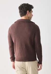 Homme avec des cheveux bouclés foncés portant un pull à côtes marron et un pantalon beige, de dos contre un fond uni clair.