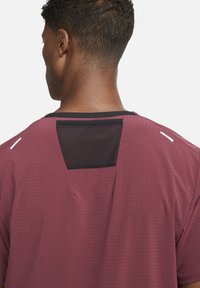 Camiseta atlética color marrón con tejido texturizado, presenta un panel de malla negra en la parte posterior y detalles blancos en los hombros.