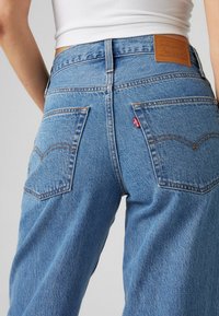 Blauwe denim jeans met een hoge taille, voorzien van twee achterzakken, een bruine leren merkpatch en contrasterende oranje stiksels op de naden.