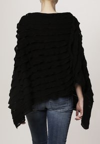 Svart stickad poncho med horisontella volanger, med en öppen design och en asymmetrisk kant. Bärs över blå jeans, vilket visar skiktad textur.