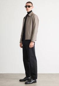 Rjav plašč s houndstooth vzorcem z zadrgo spredaj, črna polo majica, črne hlače in svetleče črne čevlje, dopolnjeno z temnimi sončnimi očali.