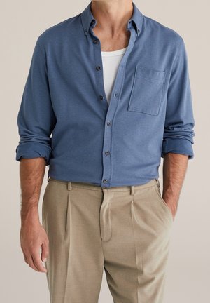Man met een blauw overhemd met knopen en opgerolde mouwen over een wit hemd en beige geplooide broek, met handen deels in de zakken.