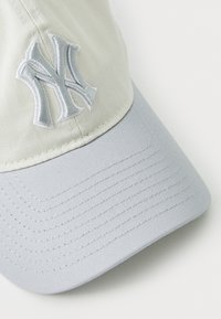Grå og lys beige kasket med buet skygge, der har et forhøjet, broderet "NY" logo på fronten. Tekstureret stof, struktureret design.