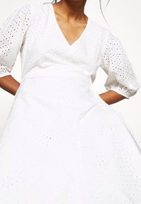 Bílé šaty s výstřihem do V, krátkými nabíranými rukávy a výšivkou průhledného materiálu. Mají stažený pas, texturovaný vzor a splývavou sukni.