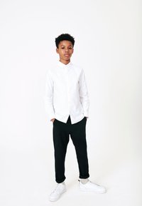 Camisa blanca abotonada con cuello, combinada con pantalones negros y zapatillas blancas. El conjunto presenta un diseño limpio y moderno.