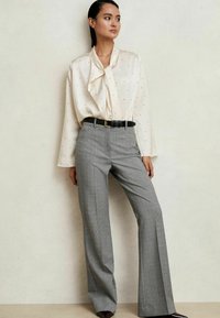 Femeie îmbrăcată cu o bluză crem cu puncte mici și un cravată, băgată în pantaloni largi cu carouri gri și o curea neagră, stând de partea unei pereți.