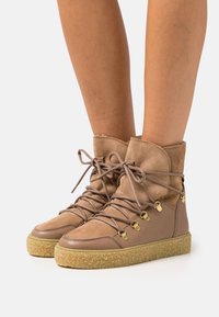 Brown suede ankle boots with a rounded toe, lace-up design, and gold eyelets. Textured rubber sole with a contrasting light brown midsole.