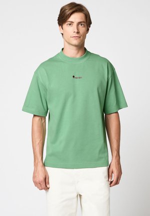 Jeune homme aux cheveux châtain clair portant un T-shirt vert à manches courtes avec un petit logo rouge et un pantalon blanc, debout devant un fond blanc uni.