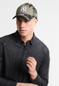 Tarnkappen-Baseballmütze mit einem weißen New York Yankees-Logo auf der Vorderseite. Material: Stoff; Farben: gedämpftes Grün, Braun und Schwarz.