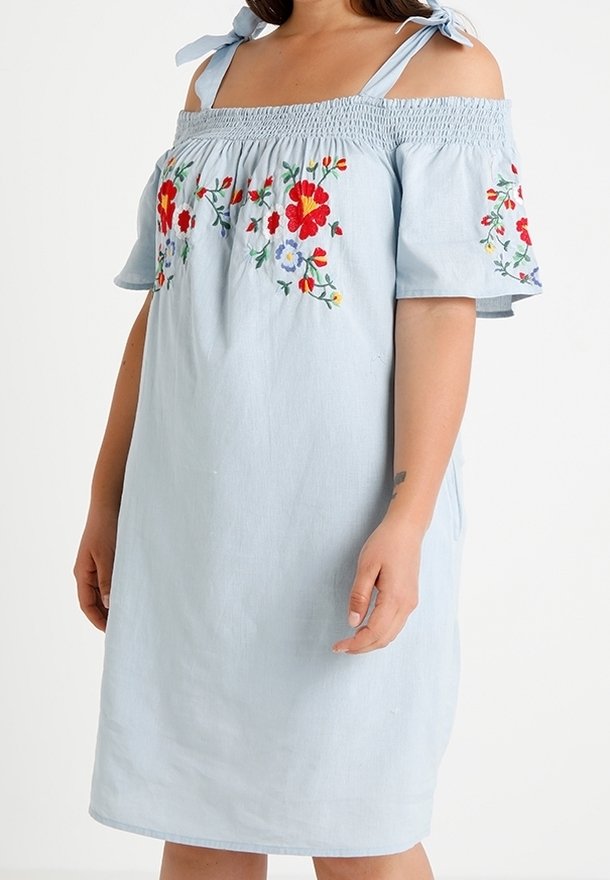 Robe bleu clair à épaules dénudées avec broderie florale rouge, orange et violette sur la poitrine et les manches, portée par une personne avec un bras relaxé.