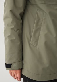 Manica di una giacca verde e tasca laterale con cinturino regolabile al polsino e logo ricamato, indossata da una persona con la mano visibile.