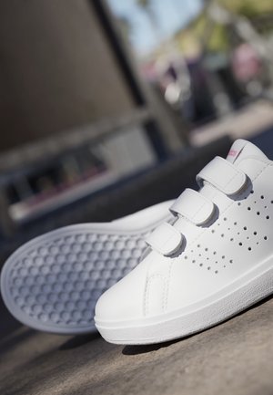 Hvide lave sneakers med tre velcrobånd og perforerede sidedetaljer, vist udendørs på en betonoverflade.
