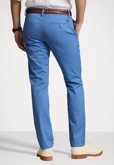 Chinos in cotone blu con un taglio dritto, caratterizzati da dettagli nelle tasche posteriori e una cintura intrecciata marrone, completati da scarpe di colore chiaro.