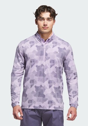 Young man wearing a lavender patterned quarter-zip pullover and dark lavender pants, standing and facing forward against white background.