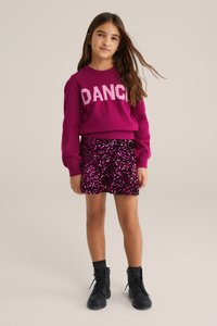 Pulover fucsia cu cuvântul "DANCE" scris în litere roz, asortat cu o fustă paietată roz și neagră, și cizme negre cu șireturi.