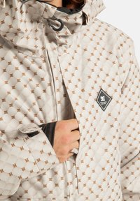 Osoba w beżowej kurtce w wzory z małymi brązowymi logo, ręka częściowo w kieszeni przedniej, czarny mankiet widoczny pod rękawem.