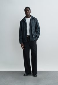 Mann steht vor einer schlichten Wand, trägt eine schwarze, gemusterte Jacke mit Reißverschluss, ein weißes T-Shirt, schwarze Hosen und schwarze Schuhe. Die Hände hängen entspannt seitlich herunter.