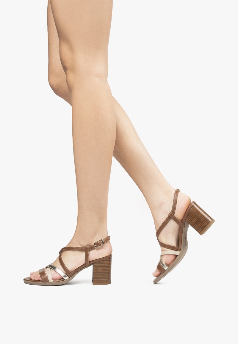 Sandales à talons brunes avec des straps croisées, présentant un accent métallique brillant, un talon en bois texturé et un design à bout ouvert.