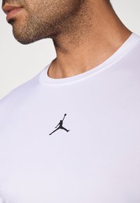 Nærbilde av en manns nedre ansikt og nakke som har på seg en hvit skjorte med en svart Jumpman-logo sentrert på brystet.