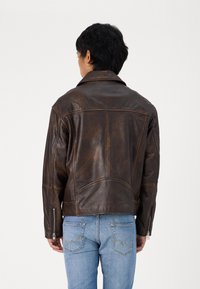 Giacca di pelle marrone con un design retrò, caratterizzata da un colletto a vista, polsini con zip e dettagli di cucitura visibili. Indossata sopra jeans blu.