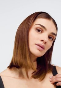 Hladké, středně dlouhé vlasy s lesklým, světle hnědým nádechem. Osoba má světlou pleť a na sobě černý top. Neutrální pozadí.