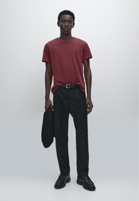 Rotes Baumwoll-T-Shirt mit kurzen Ärmeln, kombiniert mit schwarzen Hosen und schwarzen Schuhen. Hält eine schwarze Jacke und steht vor einem hellgrauen Hintergrund.