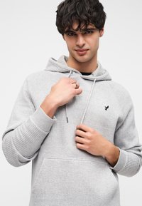 Γκρι φούτερ με κουκούλα και υφή, κορδόνι για δέσιμο και τσέπη καγκουρό, με μικρό μαύρο λογότυπο στο στήθος.