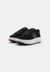 Svarta mockaskor med vit sula, rund tå och texturerade detaljer. Har svarta snören och logotypdetalj på sidan.