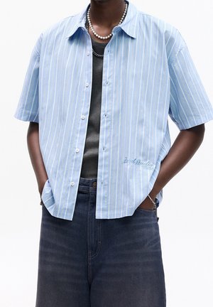 Osoba nosząca jasnoniebieską, pionowo prążkowaną koszulę z krótkim rękawem na ciemnoszarym topie, ciemne dżinsy, perłowy naszyjnik i łańcuszkową bransoletkę, ręce w kieszeniach.