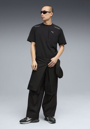 Man draagt een zwart Puma-shirt, wijde zwarte broek, zonnebril en grijze sneakers, staand tegen een grijs verloopachtergrond.