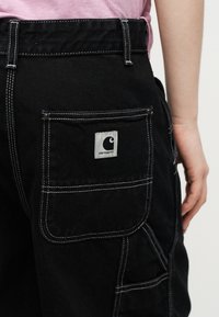 Άτομο που φοράει μαύρο παντελόνι Carhartt με άσπρα ραφές και τσέπη με το λογότυπο Carhartt, δίπλα στο χέρι του που φορά ροζ πουκάμισο.