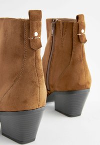 Stivali ankle in suede marrone con chiusura a zip e dettagli decorativi a cinturino, dotati di un tacco largo nero e una texture liscia.