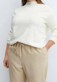 Suéter color crema con un cuello alto y mangas largas, combinado con pantalones beige con cordón. Textura suave y ajuste relajado.