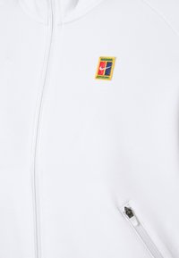 Hvid zip-up hoodie lavet af blødt materiale, med et lille flerfarvet broderet logo på brystet og en sidelukket lomme.