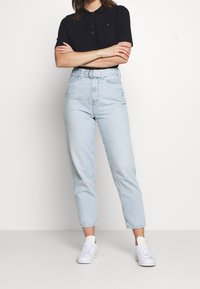 Femme portant un polo noir à manches courtes, un jean taille haute bleu clair avec une ceinture et des baskets blanches, se tenant les bras croisés.