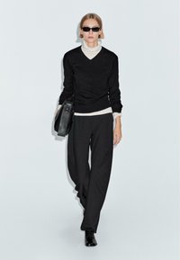 Maglione nero sopra un dolcevita bianco, abbinato a pantaloni neri a gamba larga. Borsa nera in mano e occhiali da sole completano il look. Texture morbide.