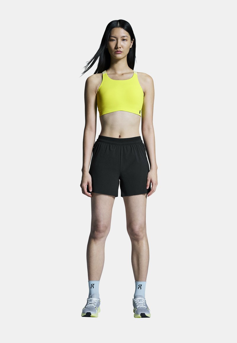 Geel sportbeha met een cropped ontwerp, gecombineerd met zwarte sportshorts. Heeft grijze schoenen met gele accenten en lichtblauwe sokken.