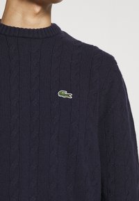 Jersey de punto de cable en color navy con cuello redondo acanalado, que presenta un pequeño logo de cocodrilo verde bordado en el pecho. Textura de punto resistente.