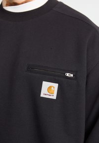 Juodas megztinis su užtrauktuku kišenėje ir baltu audinio lipduku su oranžiniu logotipu. Pagamintas iš tekstūruoto audinio, su raukšlėtu kaklu.