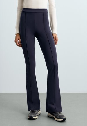 Modèle portant un pantalon bleu marine évasé à taille haute, un pull crème et des baskets grises, sur un fond de studio uni.