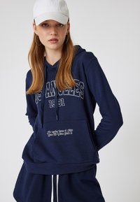 Marinblå hoodie med vit text "Los Angeles 1991" och textaccent. Har en framficka och justerbar huva med dragsko.