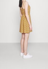 Gul blommig klänning med tunna stjärnor, rynkad rygg, utsvängd kjol och en strukturerad tyg. Buren med vita sneakers, visad från sidan.