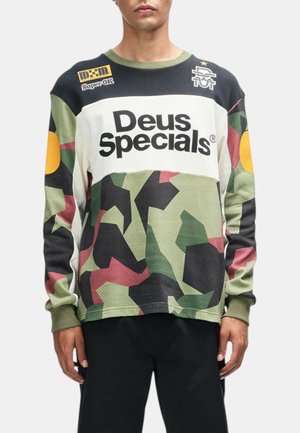 Hombre que lleva una camisa de manga larga con patrón de camuflaje en verde, negro y rojo y texto "Deus Specials" en el pecho.