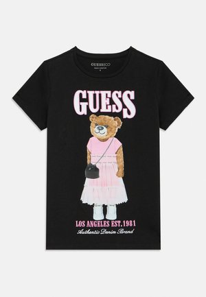 T-shirt pour enfants noirs avec un ours en peluche portant une robe rose, des chaussures blanches et tenant un petit sac noir, logo GUESS au-dessus du texte.