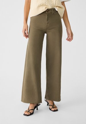 Vrouw die olijfgroene wijde broek met rafelige zomen draagt, gecombineerd met zwarte sandaaltjes met hak en bandjes en een crèmegebreid vest over een wit overhemd.