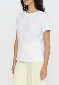 T-shirt bianca in cotone con maniche corte, caratterizzata da un piccolo dettaglio decorativo dorato sul lato sinistro del petto. Tessuto liscio, vestibilità comoda.