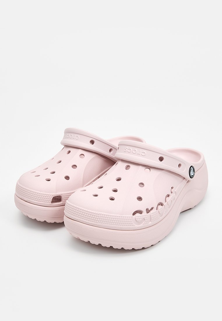 Zuecos de color rosa claro con agujeros de ventilación, una correa trasera y suelas texturizadas; presentan el logotipo "Crocs" grabado en el costado.
