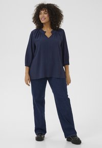 Marineblaues, strukturiertes Blusenshirt mit drei Viertelärmeln und V-Ausschnitt, kombiniert mit dunkelblauen geraden Jeans und schwarzen Schuhen.