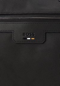 Sac en tissu noir texturé avec un accent en cuir noir comportant un logo "BOSS" en relief et des points colorés en noir, blanc et or. Fermeture zippée.