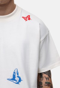 Witte T-shirt met korte mouwen, voorzien van een rood vlinderafdruk op de rechter schouder en een blauwe vlinderafdruk op de linker heup. Zacht textuur.
